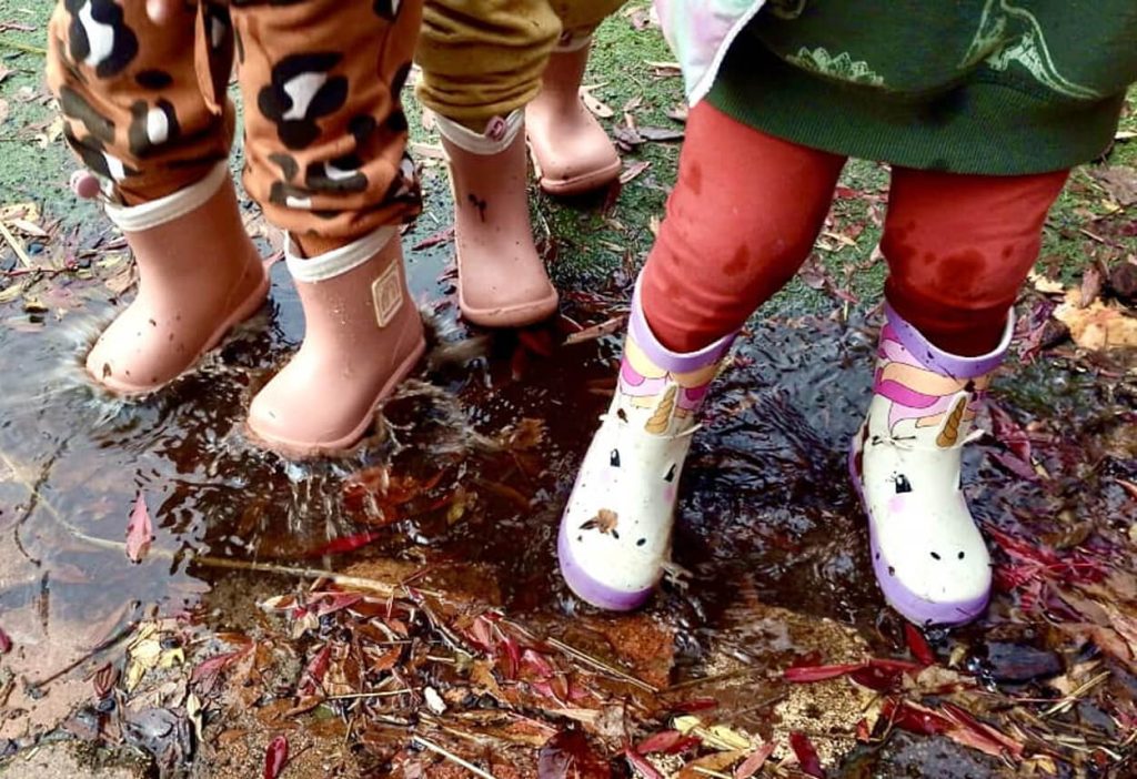Three children in rain boots stand in a muddy puddle surrounded by wet leaves and grass.