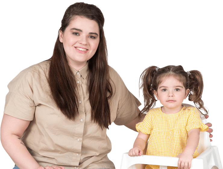 An adult woman with long brown hair sits beside a young girl in a yellow checkered dress on a white chair, sharing a moment during the Toddler Program. The woman wears a beige shirt while the girl’s hair is styled in pigtails.