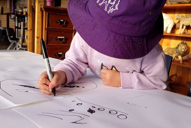 A young child wearing a purple hat draws with a black marker on large sheets of paper at a table.