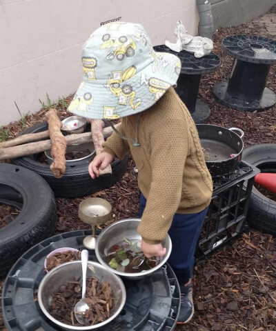 A young child wearing a patterned hat and brown sweater stands outdoors, playing with bowls filled with leaves and sticks, surrounded by tires and wooden logs.